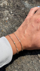 May include: Two delicate silver bracelets are worn on a wrist. One features small silver beads, the other has clear, faceted beads. The bracelets are displayed against a natural, textured background.