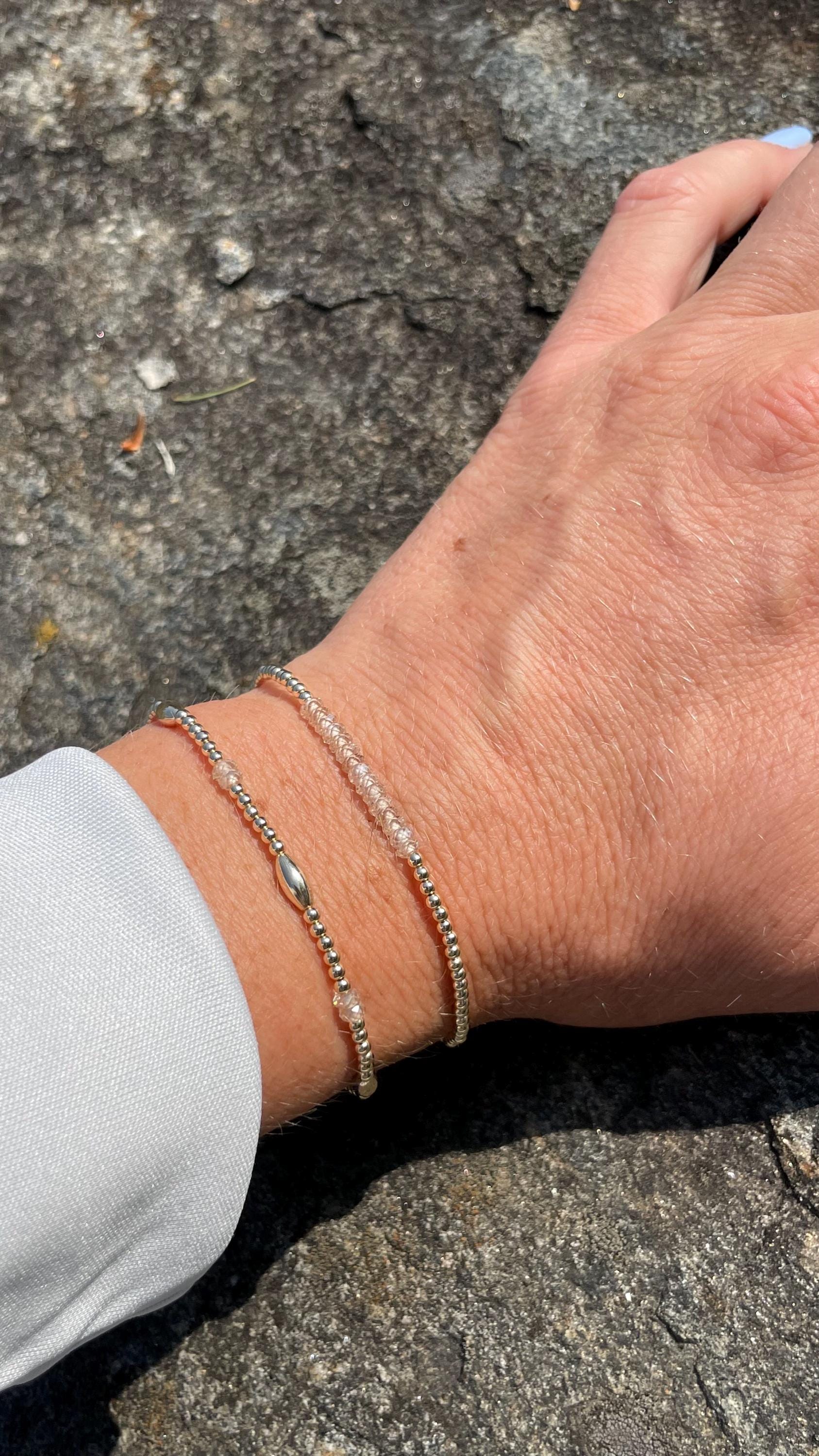 May include: Two delicate silver bracelets are worn on a wrist. One features small silver beads, the other has clear, faceted beads. The bracelets are displayed against a natural, textured background.